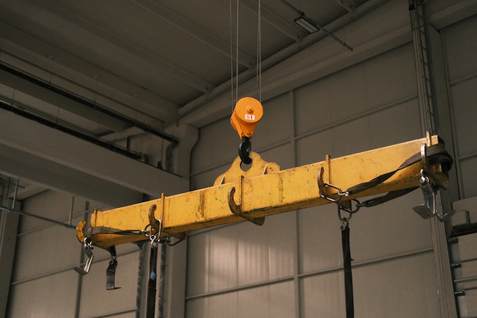 Inside an industrial warehouse, a large yellow lifting clamp with a textured, slightly worn surface is suspended from an orange hoist by a steel cable. The clamp features multiple metal hooks and secu