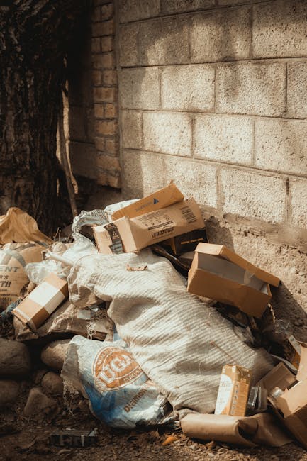 A pile of discarded cardboard boxes, some flattened and others still with their shapes intact, sits on the ground next to a rough, unfinished concrete wall and a brick wall. The boxes vary in size, wi