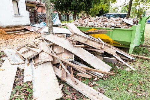 A scattered pile of weathered wooden planks, broken doors, and discarded timber of various sizes and textures lies across a grassy area next to a green skip filled with mixed rubble, bricks, and debris, in an outdoor setting near a residential building with visible windows and a partially visible staircase, suggesting recent renovation or demolition work in the vicinity, with trees and a paved pathway in the background. The scene indicates a waste clearance process, likely handled by a local rubbish removal service such as Rubbish Boy operating in the area, possibly in or around a town postcode within the UK.