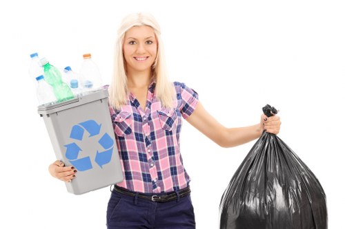 A young woman with shoulder-length blonde hair, smiling and wearing a short-sleeved, pink and purple checked shirt, stands against a plain white background. She holds a light grey recycling bin filled with various plastic bottles, including green, clear, and blue-tinted containers, with some caps visible. In her other hand, she carries a large black plastic rubbish bag, which appears to be partially full. The image emphasizes waste collection and recycling, aligning with rubbish removal services. Her posture is upright, and she extends her arms slightly outward to display the bin and the bag clearly. The scene is clean, well-lit, and professional, highlighting responsible waste disposal practices, characteristic of the services offered by companies like Rubbish Boy in the UK.