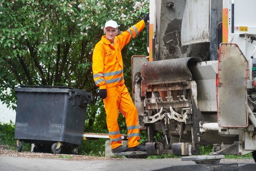 A waste collection worker dressed in high-visibility orange safety clothing, including a reflective striped jacket and trousers, is standing on a paved surface next to a large industrial waste collection vehicle. The worker is holding a small tool in his gloved hand and appears to be operating or inspecting the machinery on the side of the vehicle, which is positioned near a black plastic wheelie bin. The bin is placed on the ground and is awaiting collection, with a background of lush green trees and foliage. The scene is outdoors in a suburban or residential area, possibly in a town within the UK, as suggested by the context of rubbish removal services offered by Rubbish Boy. The vehicle's metallic surface shows signs of use, with some dirt and grime visible, and the overall environment suggests a routine waste collection operation. The lighting indicates daytime, with natural light illuminating the scene clearly, emphasizing the details of the worker's clothing, the waste vehicle, and the surrounding greenery, fitting the context of professional rubbish management services in the local area.