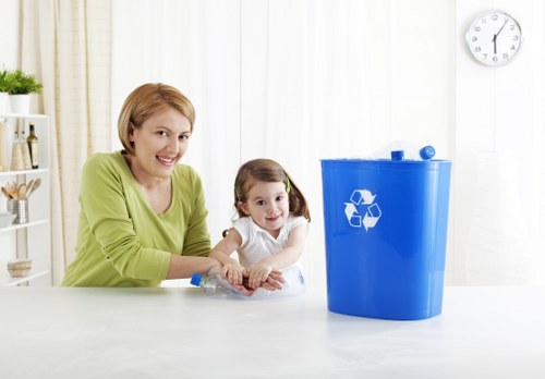 A woman with short blonde hair wearing a light green top is sitting at a white kitchen counter alongside a young girl with dark blonde hair tied in pigtails, dressed in a white top. They are both smiling and looking towards the camera. On the counter in front of them is a small, clear plastic container, and to the right, there is a large, blue plastic recycling bin with a white recycling symbol printed on it, positioned in a bright, modern kitchen. The background features white vertical blinds covering windows, a white wall with a round clock showing approximately 12:15, and a white shelving unit with decorative items including a small potted plant and a glass jar. The scene is well-lit with natural daylight, emphasizing the clean and organized environment, and subtly relates to waste management and recycling services provided by Rubbish Boy.